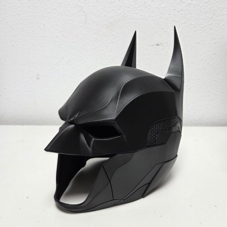 May include: A black Batman cowl, featuring a pointed nose, eye openings, and tall, pointed ears. The mask has a textured surface and is designed to fit the head. The overall design is sleek and angular.