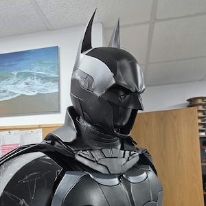May include: A black and gray Batman costume, featuring a cowl with pointed ears, a chest emblem, and armored plating. The costume is detailed with silver accents and a cape, showcasing a superhero theme.