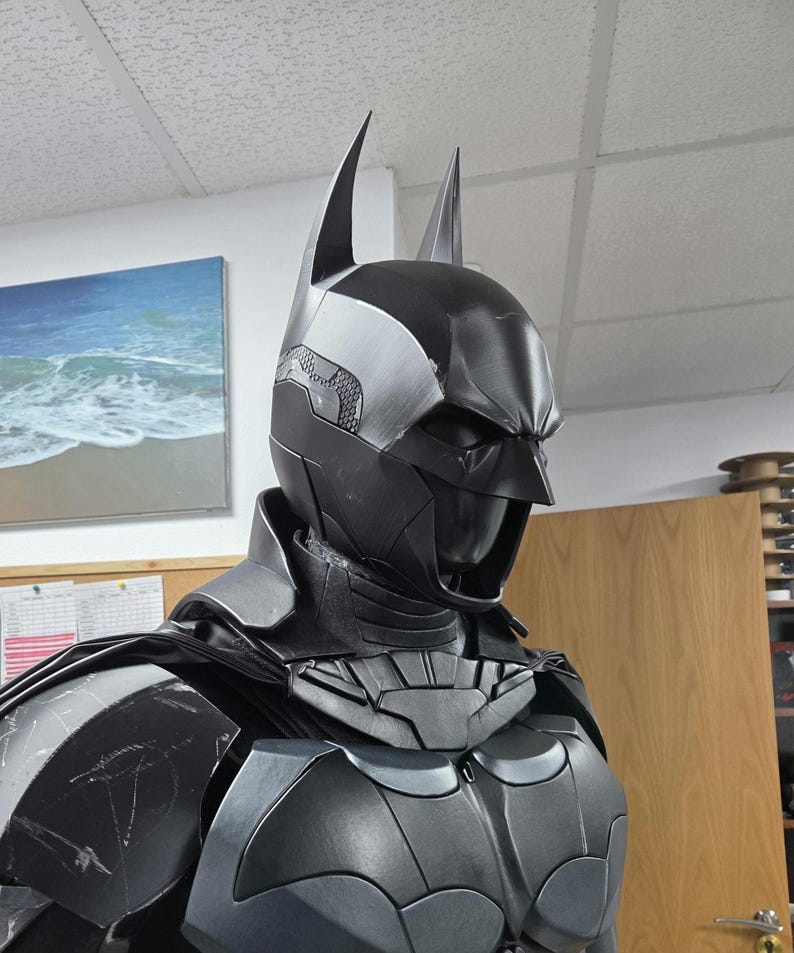 May include: A black and gray Batman costume, featuring a cowl with pointed ears, a chest emblem, and armored plating. The costume is detailed with silver accents and a cape, showcasing a superhero theme.