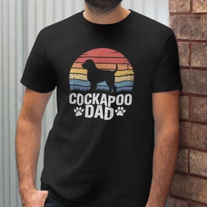 Puede incluir: Camiseta negra con la silueta de un perro Cockapoo sobre un diseño de puesta de sol. El texto "Cockapoo Dad" es blanco, con huellas de patas. La camiseta está hecha de un material suave.