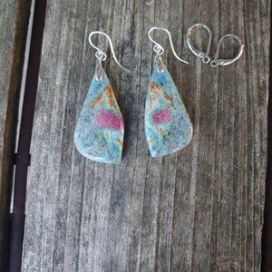 May include: A pair of silver earrings with teardrop-shaped blue and pink gemstone drops. The gemstones have a marbled pattern and are set in silver.