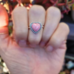 May include: A silver ring with a pink heart-shaped gemstone. The ring is on a finger.