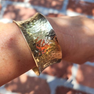 May include: A gold hammered metal cuff bracelet. The bracelet is wide and has a rounded shape. It is made of a textured metal.