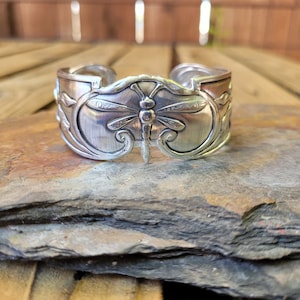 May include: A silver bracelet featuring a dragonfly design. The bracelet is crafted from a recycled spoon and has a hammered finish.