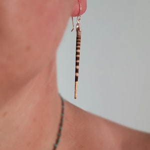 May include: A pair of silver dangle earrings with a long, thin, striped brown and white feather pendant with a gold tip.