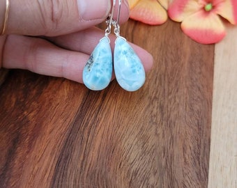 Könnte beinhalten: Ein Paar tropfenförmige Ohrringe mit hellblauem und weißem Marmormuster. Die Ohrringe sind an silberfarbenen Klappbügel-Ohrdrähten befestigt. Der Hintergrund umfasst eine Holzoberfläche und orangefarbene Blumen.