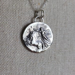 Puede incluir: Un collar con un colgante de plata que presenta un relieve detallado de una mujer montando un caballo alado. El colgante es redondo y tiene una superficie texturizada.