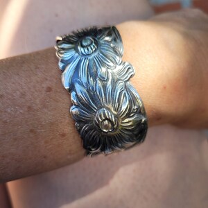 May include: A silver bracelet with a floral design. The bracelet is made of metal and has a detailed design of three flowers.