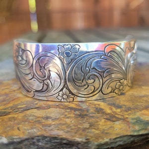 May include: A wide, silver cuff bracelet with an engraved floral and scroll design. The cuff is made of polished metal and features detailed carvings of flowers and swirling patterns. The bracelet is displayed on a natural stone surface.