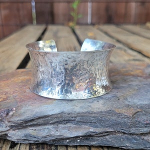 May include: A wide, hammered silver cuff bracelet. The metal has a textured, dimpled surface. The bracelet has an open design and is displayed on a grey stone surface.