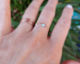 May include: A delicate gold chain ring with a single light blue gemstone. The ring is worn on a finger.