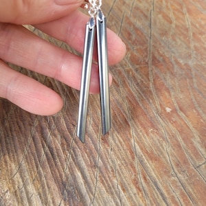 May include: A pair of silver-colored, linear dangle earrings. Each earring features a long, slender, rectangular bar with a pointed tip. The earrings are suspended from a small, decorative silver hook. The background is a textured, wooden surface.