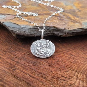 Può includere: Una collana d'argento con un ciondolo rotondo che presenta una rappresentazione dettagliata di due figure in una scena. Il ciondolo è su una catena d'argento.