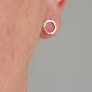 May include: A small, circular stud earring with a metallic gold finish and a textured, hammered surface. The earring features an open center design and is worn on the earlobe, showcasing a reflective, irregular edge that catches the light.