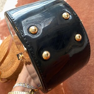 May include: A black patent leather handbag with a glossy finish, featuring four gold-tone metal feet on the bottom panel. The bag has a structured, rounded shape with visible stitching along the edges and gold-tone hardware accents.