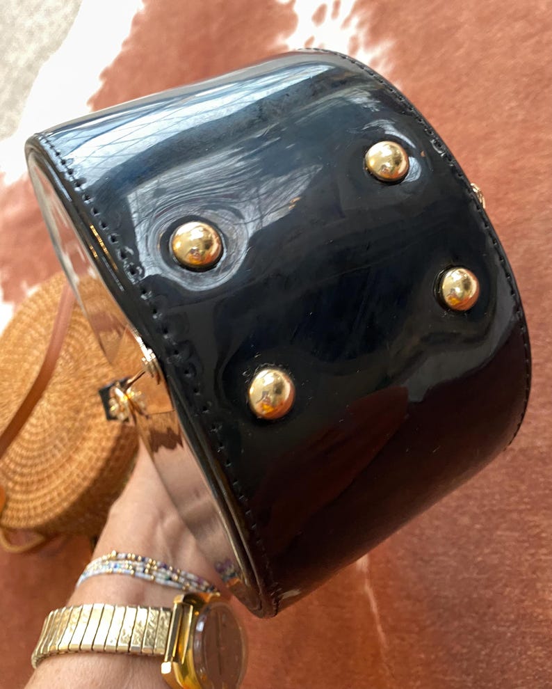May include: A black patent leather handbag with a glossy finish, featuring four gold-tone metal feet on the bottom panel. The bag has a structured, rounded shape with visible stitching along the edges and gold-tone hardware accents.