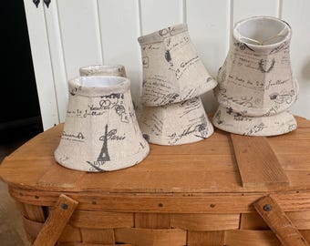 May include: A set of four small lamp shades made of beige fabric with a black printed pattern featuring the Eiffel Tower, cursive French script, and various postal-style stamps. Each shade has a tapered shape and a white interior lining.