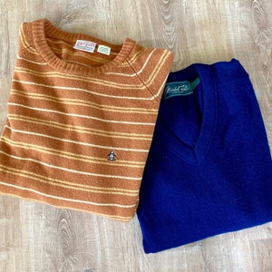May include: Two sweaters, one brown with white stripes and a penguin logo, and one navy blue with a v-neck. The brown sweater has the text "penguin" on the tag. The navy blue sweater has the text "Marshall Field's" on the tag.