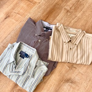 Puede incluir: Tres camisas abotonadas con rayas verticales. Una es azul claro con rayas blancas, otra marrón con rayas burdeos y la tercera beige con rayas marrones. Las camisas están dobladas sobre una superficie de madera.