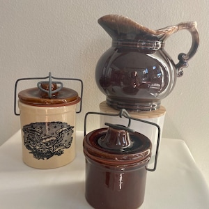 Könnte beinhalten: Eine Sammlung von Vintage-Keramik-Küchenutensilien. Enthält einen dunkelbraunen Krug mit Henkel, zwei kleine Gläser mit Deckel und Drahtgriffen sowie ein beiges Glas mit schwarzem Adler-Motiv. Die Artikel sind auf einer weißen Oberfläche angeordnet.