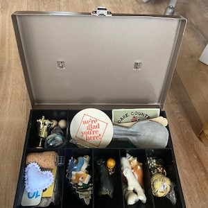 Puede incluir: Una caja abierta, de color metálico, dividida en compartimentos, llena de varios objetos pequeños. Incluye un trofeo dorado, un corcho, un corazón de fieltro, un posavasos con texto, una pequeña pala y varias figuritas.