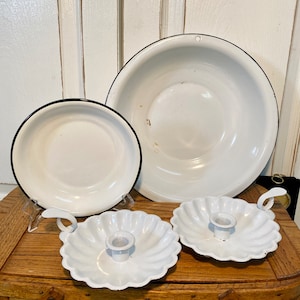 Puede incluir: Una colección de artículos de cocina de esmalte blanco, que incluye dos platos, un cuenco grande y dos candelabros festoneados. Cada pieza tiene un borde negro. Los artículos están dispuestos sobre una superficie de madera.