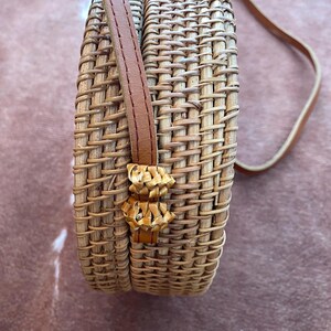 May include: A round, woven rattan crossbody bag featuring a light tan natural finish. The bag has a structured cylindrical shape with a vertical weave pattern and a thin, smooth brown leather shoulder strap attached to the side with woven loops.