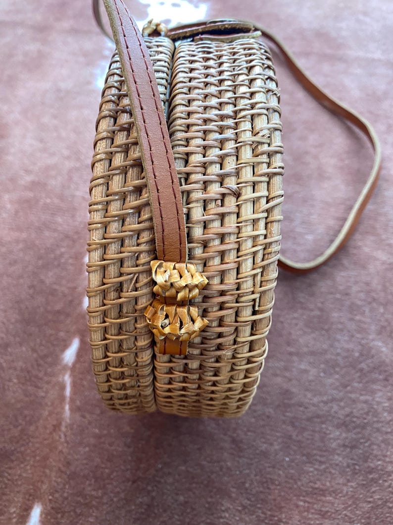 May include: A round, woven rattan crossbody bag featuring a light tan natural finish. The bag has a structured cylindrical shape with a vertical weave pattern and a thin, smooth brown leather shoulder strap attached to the side with woven loops.
