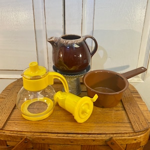 Könnte beinhalten: Eine Sammlung von drei Vintage-Teekannen. Eine ist braun mit einer glasierten Oberfläche, eine ist aus klarem Glas mit einem gelben Deckel und Griff und die andere ist aus gelbem Kunststoff mit einem Griff und einem Ausguss.