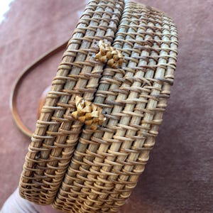 May include: A round, structured crossbody bag crafted from woven light brown rattan material. The bag features a secure lid with two small, braided rattan loops acting as a closure mechanism and a thin, matching brown leather shoulder strap.