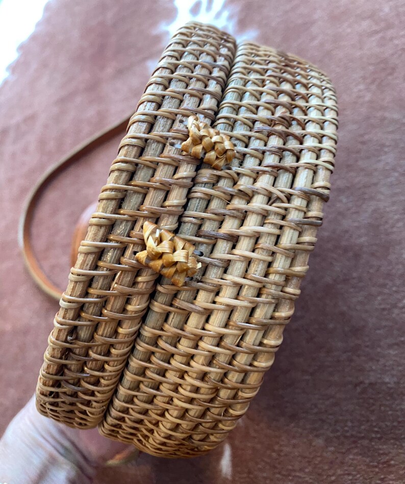 May include: A round, structured crossbody bag crafted from woven light brown rattan material. The bag features a secure lid with two small, braided rattan loops acting as a closure mechanism and a thin, matching brown leather shoulder strap.