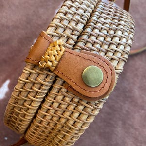 May include: A round woven rattan crossbody bag featuring a light brown natural finish. The bag includes a tan leather strap and a matching leather closure tab secured with a circular brass-toned metal snap button fastener on the front.