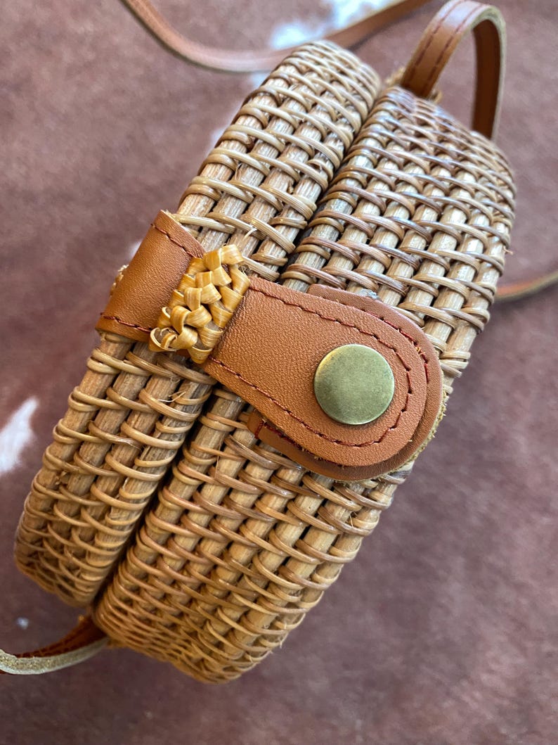 May include: A round woven rattan crossbody bag featuring a light brown natural finish. The bag includes a tan leather strap and a matching leather closure tab secured with a circular brass-toned metal snap button fastener on the front.