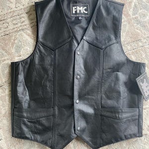 May include: A black leather waistcoat featuring a V-neckline, four front snap button closures, and two lower pockets with flaps. The interior label displays the text FMC and the size 46. A matching brand tag is attached to the side of the garment.