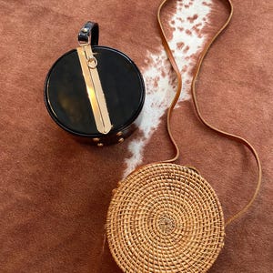 May include: A round black handbag with a glossy finish, a top handle, and a gold-tone metal clasp, paired with a round woven rattan crossbody bag featuring a long brown leather strap. Both bags are circular in shape and display distinct textures.