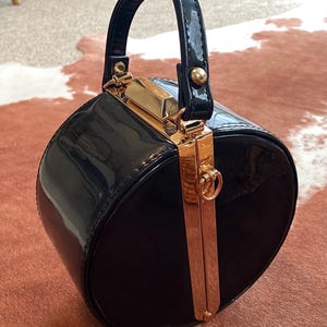 May include: A round, black patent leather handbag with a glossy finish. The bag features a structured top handle and a vertical gold-toned metal frame closure that runs down the centre, complete with a small circular ring detail at the top.