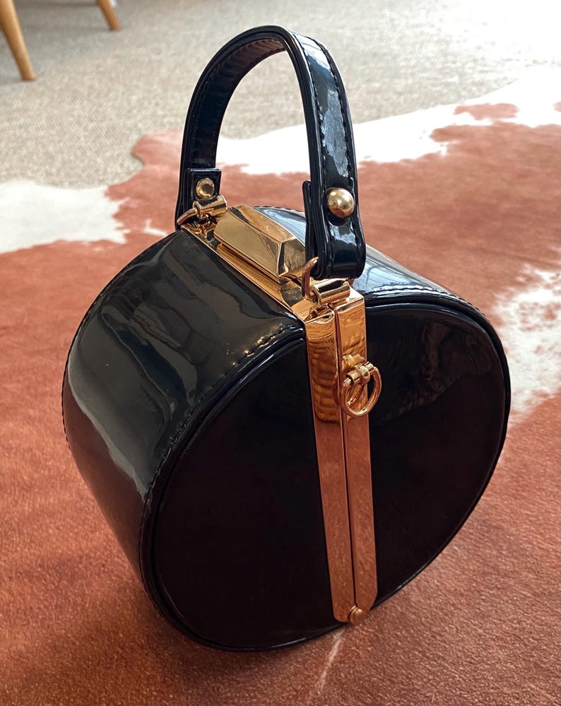 May include: A round, black patent leather handbag with a glossy finish. The bag features a structured top handle and a vertical gold-toned metal frame closure that runs down the centre, complete with a small circular ring detail at the top.