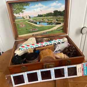 Puede incluir: Caja de madera abierta con tapa con bisagras, que muestra una escena de postal antigua de un parque. En el interior hay varios objetos, incluyendo un búho de madera, pajitas decorativas y rollos de cinta. Varios contenedores de diapositivas están al frente.