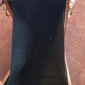 May include: A rectangular black clutch bag with a glossy finish and a gold-toned metal frame. The interior is lined with black fabric. The bag features gold-toned metal hardware, including side rings for a strap and a top clasp closure mechanism.