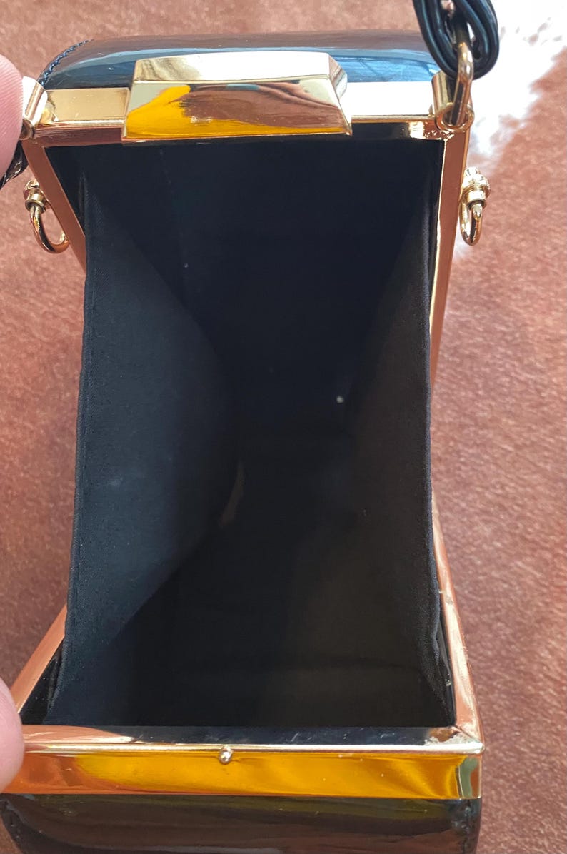 May include: A rectangular black clutch bag with a glossy finish and a gold-toned metal frame. The interior is lined with black fabric. The bag features gold-toned metal hardware, including side rings for a strap and a top clasp closure mechanism.