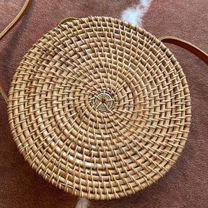May include: A round, woven rattan crossbody bag featuring a natural light brown finish with a spiral pattern radiating from the centre. The bag includes a thin, adjustable brown leather strap attached to the sides for carrying over the shoulder.