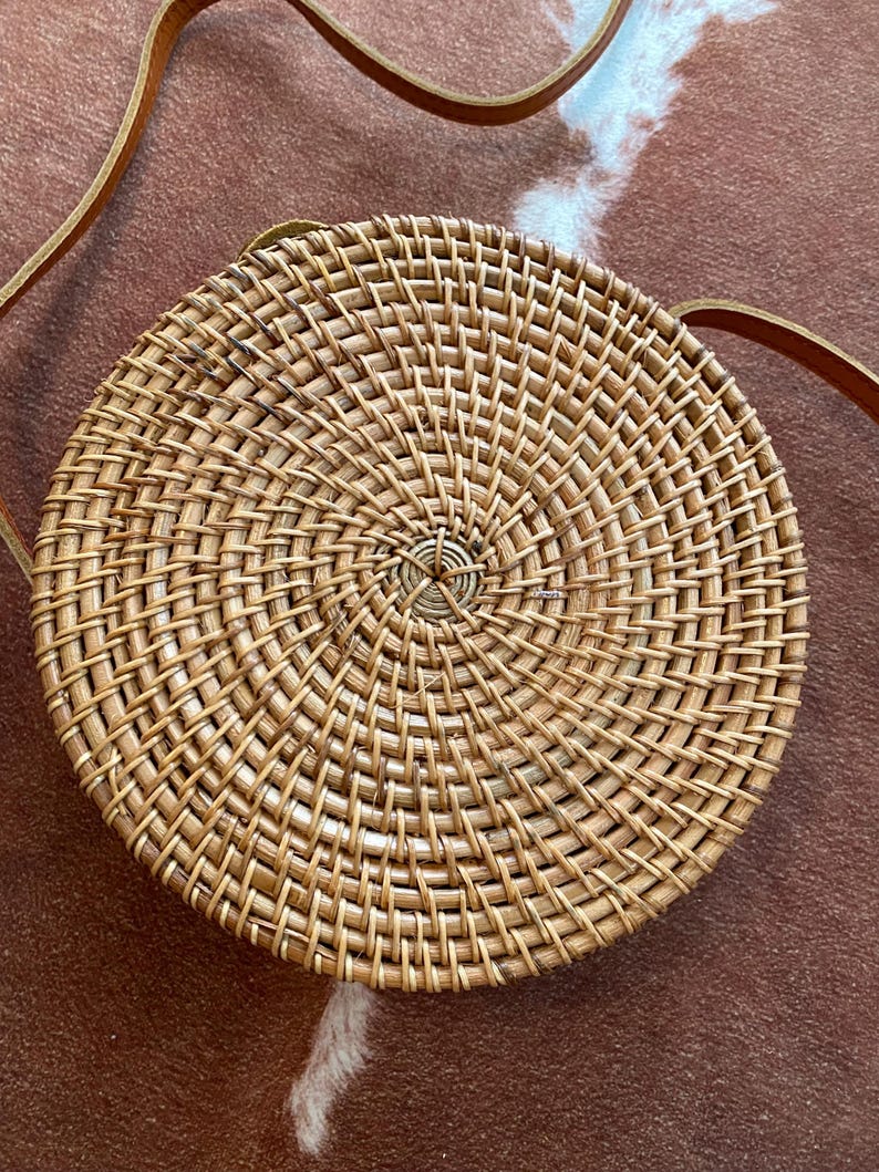 May include: A round, woven rattan crossbody bag featuring a natural light brown finish with a spiral pattern radiating from the centre. The bag includes a thin, adjustable brown leather strap attached to the sides for carrying over the shoulder.