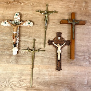 May include: Five different crucifixes with various styles and materials. The crucifixes are made of wood, metal, and seashells. They are all hanging on a wooden wall.
