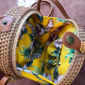May include: A round woven rattan crossbody bag with a tan finish and brown leather straps. The interior is lined with a bright yellow fabric featuring a tropical floral pattern in shades of green, white, and brown, secured by a metal snap closure.