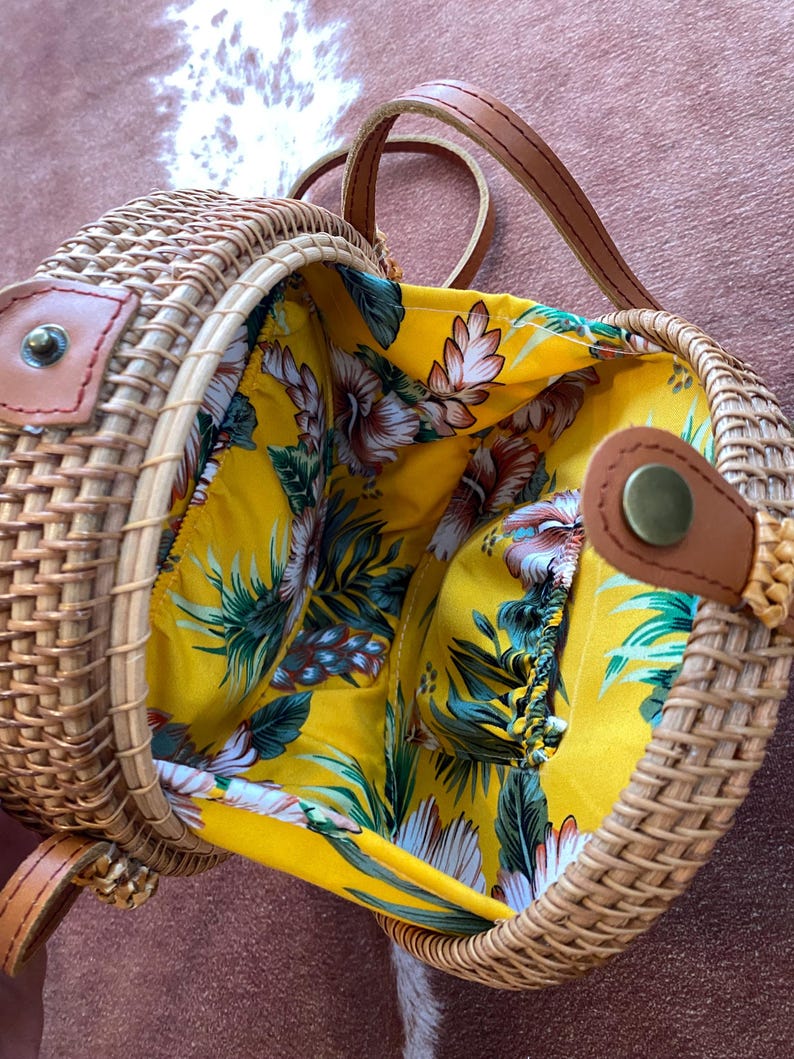 May include: A round woven rattan crossbody bag with a tan finish and brown leather straps. The interior is lined with a bright yellow fabric featuring a tropical floral pattern in shades of green, white, and brown, secured by a metal snap closure.