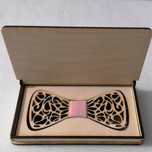 Peut inclure: Une boîte en bois avec un couvercle à charnière, ouverte pour révéler un nœud papillon en bois décoratif avec un insert en tissu rose. Le nœud papillon a un design détaillé et complexe.