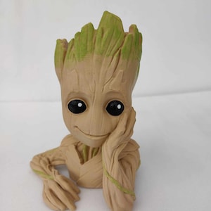 Peut inclure: Un pot en céramique brun et vert en forme de personnage du film Les Gardiens de la Galaxie. Le personnage a une tête verte feuillue et un visage souriant avec de grands yeux. Le pot repose le menton sur sa main.