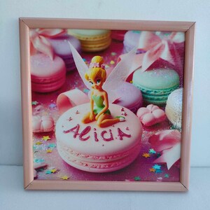 Peut inclure: Une image encadrée de rose avec une figurine de Clochette assise sur un gâteau rose et blanc avec le nom "Alicia" écrit en paillettes roses. Le gâteau est entouré de macarons et de confettis roses et blancs.