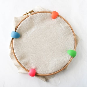 May include: A wooden embroidery hoop with beige fabric stretched inside. The hoop is secured with colorful, silicone hoop grippers in blue, orange, green, and pink. The fabric is a neutral color, and the hoop is made of wood.