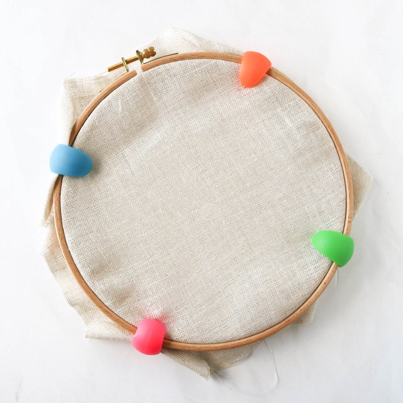 May include: A wooden embroidery hoop with beige fabric stretched inside. The hoop is secured with colorful, silicone hoop grippers in blue, orange, green, and pink. The fabric is a neutral color, and the hoop is made of wood.